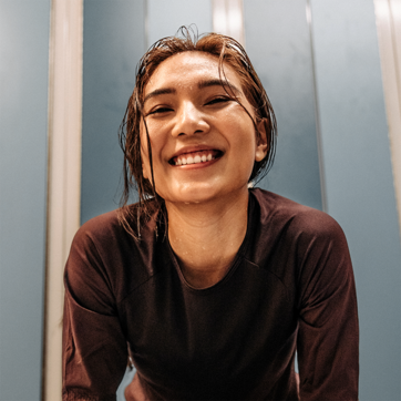 Jovem mulher sorridente com cabelo molhado, usando uma camiseta de manga longa. Ela está em um ambiente interno, aparentando estar feliz e satisfeita, com o fundo mostrando paredes em tons de azul. A imagem transmite uma sensação de alegria e energia positiva.