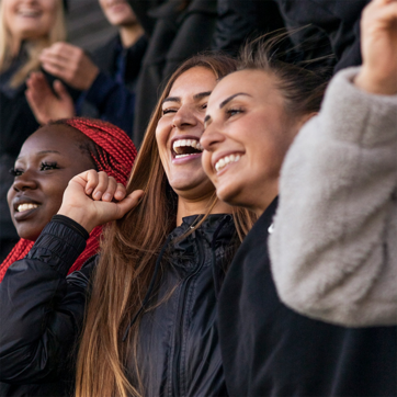 Grupo de mulheres sorrindo e se divertindo, exibindo expressões de alegria. Algumas estão levantando as mãos e a cabeça, celebrando em um ambiente animado, possivelmente em um evento esportivo ou uma apresentação. O clima é de entusiasmo e camaradagem.