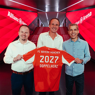 Três homens posam juntos em um corredor da Allianz Arena, segurando uma camisa laranja do FC Bayern München. A camiseta tem "2027" e "DOPPELHERZ" estampados nas costas. O fundo é iluminado com luzes vermelhas e o nome da arena é visível na parede.