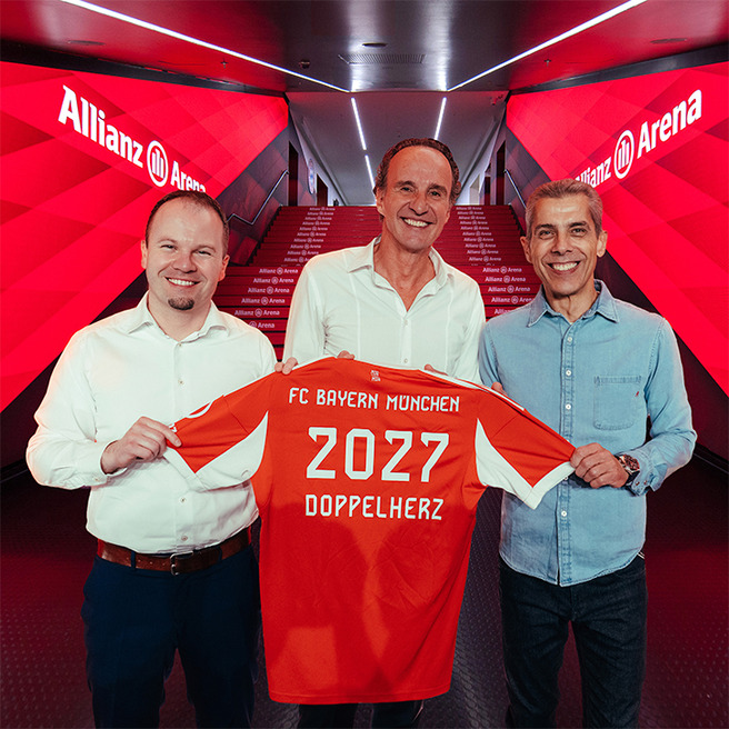 Três homens posam juntos em um corredor da Allianz Arena, segurando uma camisa laranja do FC Bayern München. A camiseta tem "2027" e "DOPPELHERZ" estampados nas costas. O fundo é iluminado com luzes vermelhas e o nome da arena é visível na parede.