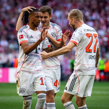 Jogadores do Bayern de Munique comemoram após um gol. Um deles, com cabelo trançado, recebe abraços dos colegas, que demonstram alegria e celebração. Todos vestem uniformes brancos e vermelhos do time, com símbolos da Bundesliga. O ambiente é de festa em um estádio lotado.